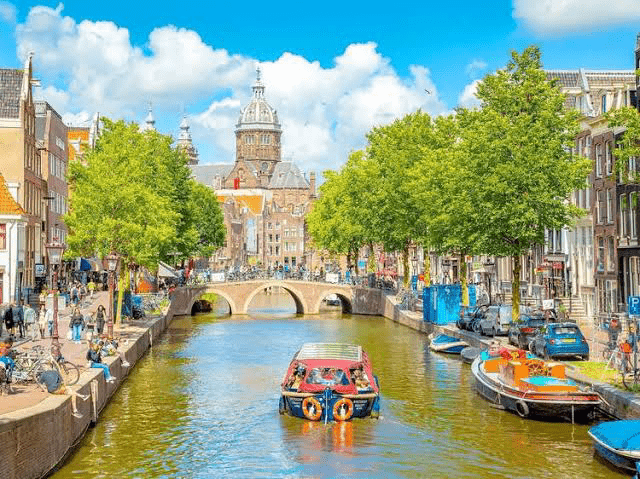 Scenic canal view in Amsterdam, showcasing a boat with life rings, lush trees, historic architecture, and a bridge under a bright blue sky.