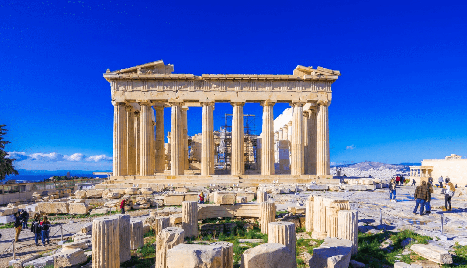 The Parthenon stands majestically atop the Acropolis in Athens, surrounded by ancient ruins. Tourists explore the site under a clear blue sky.