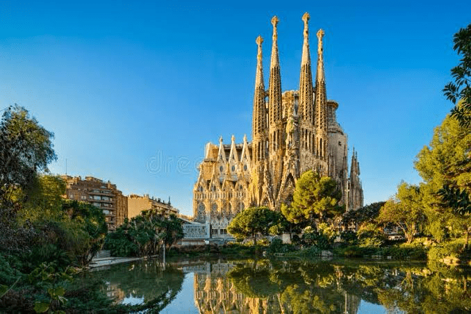 Sagrada Família, a monumental basilica in Barcelona, showcases intricate Gothic architecture reflected in a tranquil pond under a clear blue sky.