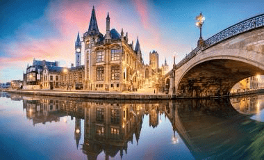 Historic buildings reflect in calm water at dusk, with a bridge arching over the river, showcasing a scenic European cityscape.
