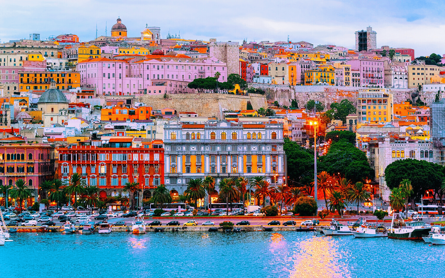 Colorful buildings line the hillside of a coastal city at dusk, reflecting vibrant hues in the water and creating a picturesque urban waterfront scene.