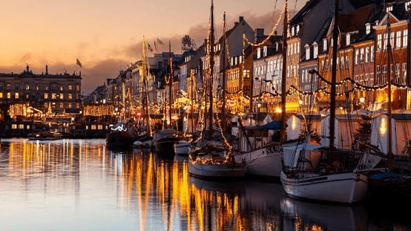Colorful buildings along a canal are illuminated by festive lights at dusk, with moored boats reflecting in the calm water.