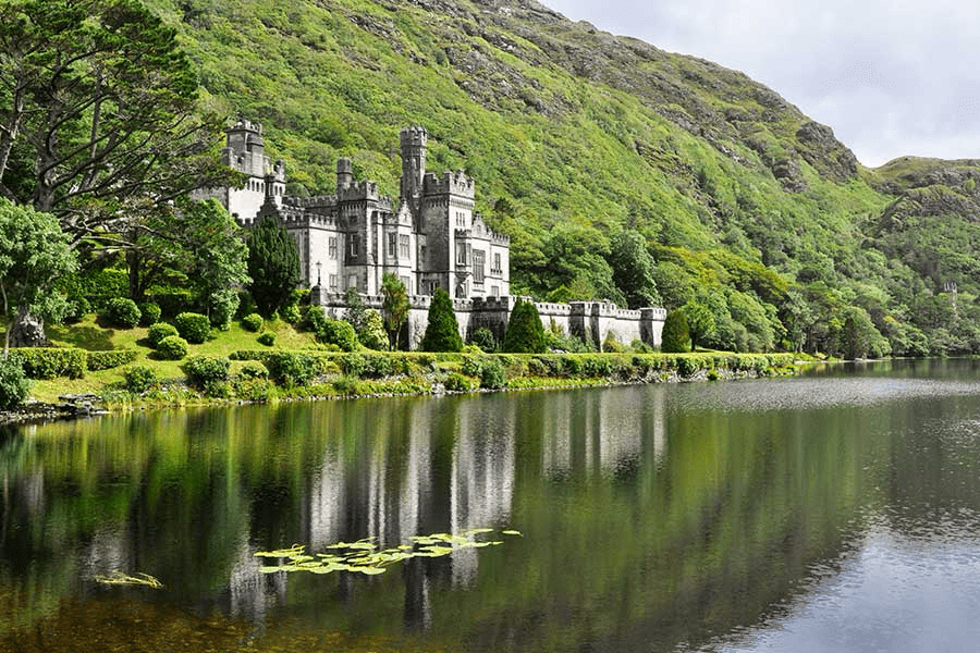 A medieval castle sits by a serene lake, surrounded by lush greenery and mountains, reflecting the scenic beauty of the landscape.