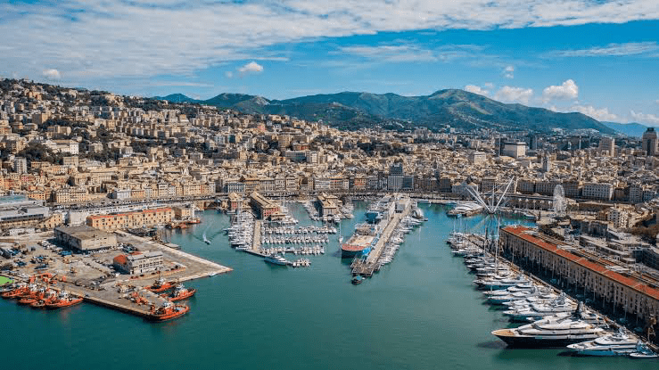 Vibrant aerial view of Genoa’s harbor, showcasing a bustling marina filled with boats and yachts, framed by colorful hillside buildings and mountains.