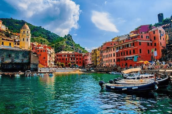 Colorful buildings line the waterfront of a coastal village, reflecting in the clear blue water. Boats are anchored, set against a backdrop of hills and a blue sky.