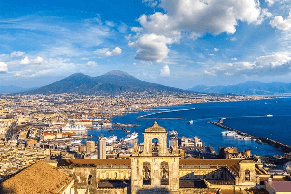 Panoramic view of Naples, Italy, featuring Mount Vesuvius in the background, the bustling harbor, and historical architecture in the foreground.