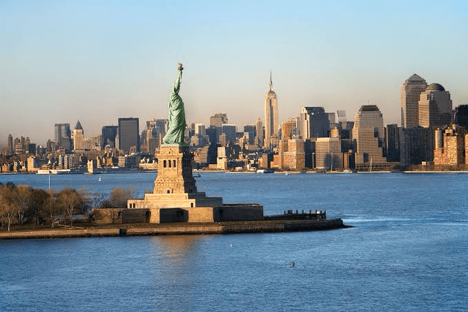 The Statue of Liberty stands prominently in the foreground, with the New York City skyline including the Empire State Building in the background.