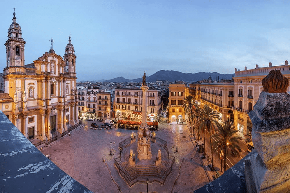 Historic square at twilight, featuring a prominent statue and illuminated baroque architecture. Palm trees and surrounding buildings enhance the vibrant atmosphere.
