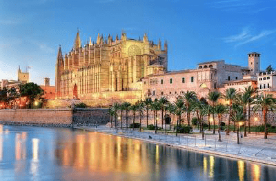 Palma Cathedral stands majestically by the waterfront, illuminated at dusk. Palm trees line the promenade, highlighting Mallorca's cultural heritage.
