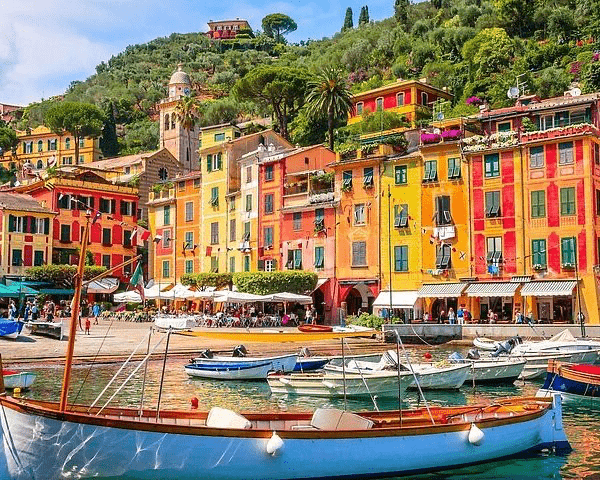 Colorful coastal buildings line the harbor of a picturesque Italian village, with boats moored in clear waters and a lush hillside in the background.