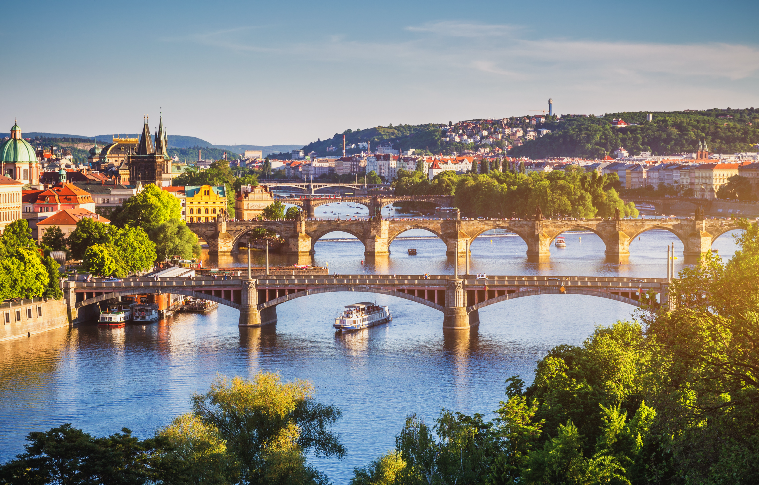 Prague's iconic bridges span the Vltava River, with a riverboat cruising beneath a backdrop of historic architecture and lush greenery.