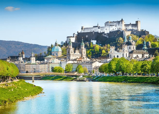 Salzburg's historic skyline features Hohensalzburg Fortress on a hill, with lush greenery and the Salzach River flowing through the foreground.