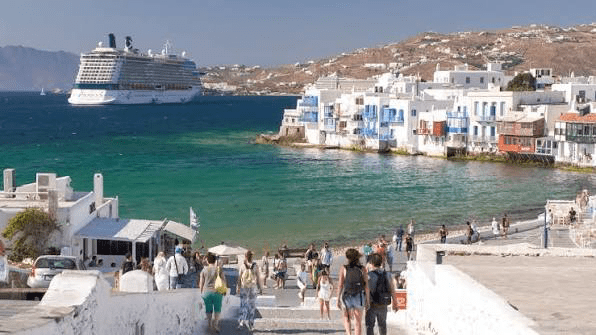 People stroll along a scenic waterfront in Mykonos, Greece, with a large cruise ship docked in the background and colorful buildings lining the shore.