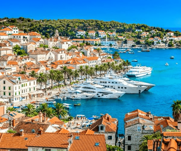 A vibrant coastal town with terracotta-roofed buildings and palm trees lining the harbor, featuring luxury yachts in turquoise waters under a clear blue sky.