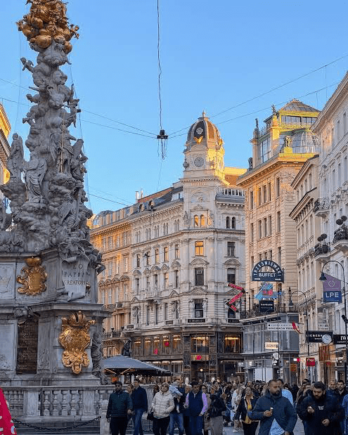 Bustling city square in Vienna featuring an ornate statue, historic buildings, and pedestrians, showcasing the city's architectural charm and vibrancy.