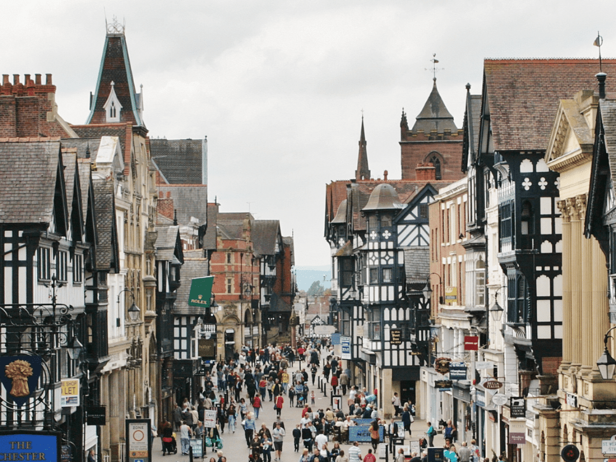 Busy street scene in a historic town, lined with traditional black-and-white buildings. Crowds of people stroll past shops and cafes under a cloudy sky.