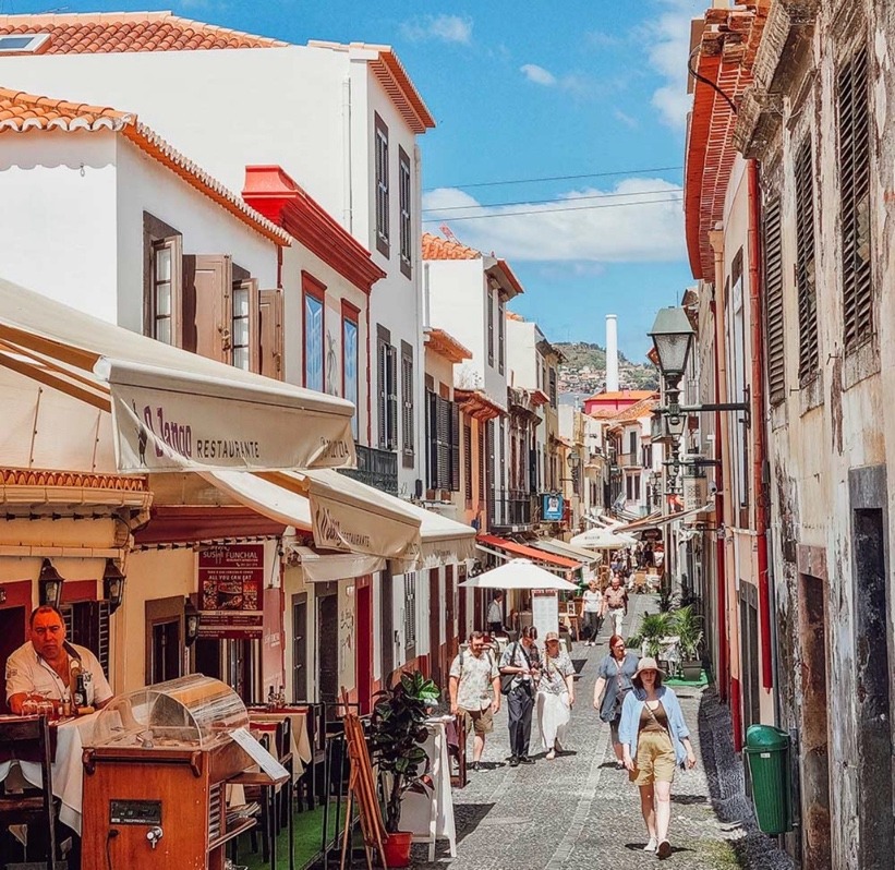 Quaint street bustling with pedestrians, lined by charming restaurants and historic buildings under a bright blue sky.