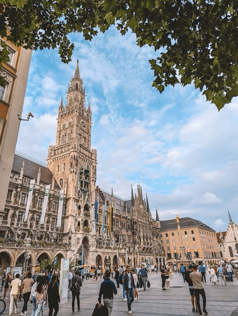 Neues Rathaus in Munich, a grand Gothic building with a towering spire, surrounded by pedestrians and vibrant banners under a partly cloudy sky.