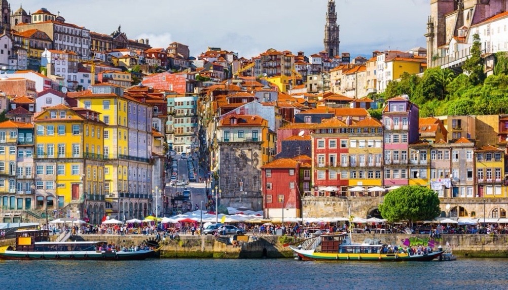 Colorful terraced buildings line the waterfront of Porto, Portugal, with bustling markets and boats along the Douro River, showcasing the city's vibrant culture.
